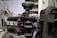 The image depicts a vintage engine in a workshop setting, with visible components such as wires, tubes, and metal parts. The surrounding environment includes shelving and diagrams on the walls, suggesting a repair or restoration area.