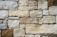 A wall composed of irregularly shaped stones stacked in a precise arrangement. The stones vary in shades of beige, off-white, and light brown, displaying a rough texture and natural appearance.