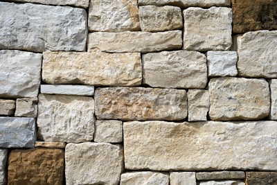 A wall composed of irregularly shaped stones stacked in a precise arrangement. The stones vary in shades of beige, off-white, and light brown, displaying a rough texture and natural appearance.