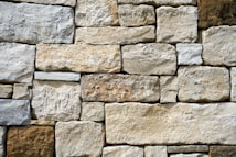 A wall composed of irregularly shaped stones stacked in a precise arrangement. The stones vary in shades of beige, off-white, and light brown, displaying a rough texture and natural appearance.