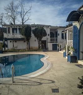 A serene outdoor scene features an empty swimming pool surrounded by a tiled deck. The pool has a metal ladder and is positioned in front of a two-story white house with blue accents, including shutters and doors. Trees and plants are present around the area, with some leafless trees visible. The sky is partly cloudy.