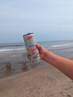 A hand holding a can of Celsius energy drink with 'Tropical Vibe' branding is positioned in the foreground against a beach backdrop. The sandy shore and gentle waves of the ocean create a relaxed seaside setting.