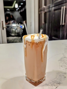 A tall glass filled with a creamy iced coffee beverage is topped with a generous amount of foam and drizzled with caramel sauce. Two straws are placed into the drink. The glass is set on a white marble countertop, and in the background, there are dark cabinets and a reflective glass door.