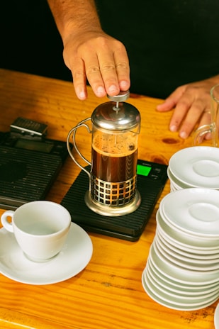 Hands pressing down a French press filled with rich, dark coffee in a cozy kitchen setting.