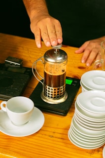 A hand pressing down on a French press filled with coffee on a wooden table. Beside it, there is a white coffee cup sitting on a saucer and a neatly stacked pile of similar saucers. A measuring scale is underneath the French press.