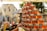 A market stall is displaying a pyramid of neatly stacked glass jars filled with golden honey, reflecting the warm sunlight. The background shows a bustling outdoor market scene, with blurred figures of people and various stalls under a clear sky.