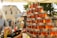 A market stall is displaying a pyramid of neatly stacked glass jars filled with golden honey, reflecting the warm sunlight. The background shows a bustling outdoor market scene, with blurred figures of people and various stalls under a clear sky.