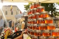 A market stall is displaying a pyramid of neatly stacked glass jars filled with golden honey, reflecting the warm sunlight. The background shows a bustling outdoor market scene, with blurred figures of people and various stalls under a clear sky.