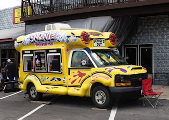 A colorful, yellow ice cream truck is parked on a street, featuring bright designs and the words 'Snowie Shaved Ice' and 'School's Out' on the top. The truck is decorated with images of shaved ice and playful patterns in purple and pink. It is situated next to a building with a wooden balcony and corrugated metal siding. There is a red folding chair beside the truck and a small group of people in the background.