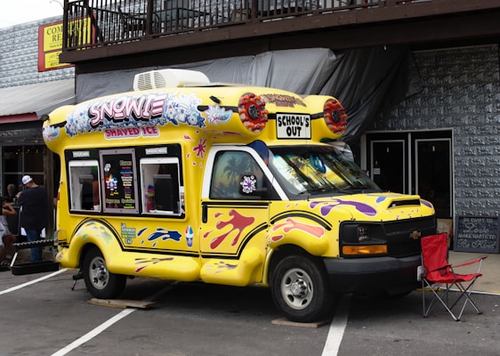 A colorful, yellow ice cream truck is parked on a street, featuring bright designs and the words 'Snowie Shaved Ice' and 'School's Out' on the top. The truck is decorated with images of shaved ice and playful patterns in purple and pink. It is situated next to a building with a wooden balcony and corrugated metal siding. There is a red folding chair beside the truck and a small group of people in the background.