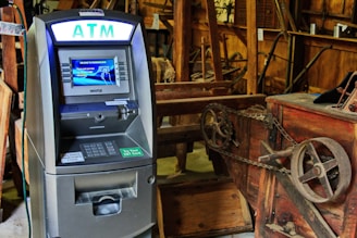An ATM situated indoors among various wooden and metal antique machinery, creating a juxtaposition between modern technology and historical equipment.