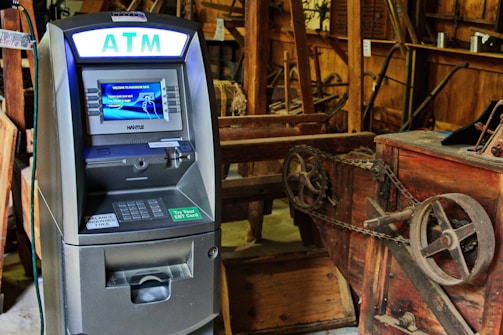 An ATM situated indoors among various wooden and metal antique machinery, creating a juxtaposition between modern technology and historical equipment.