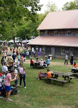 A lively outdoor gathering with numerous people of various ages engaging in activities. The scene includes a rustic wooden building with a metal roof, surrounded by picnic tables where attendees are seated. A group is in line, while others are scattered around the vicinity, possibly part of a fair or festival. Trees and greenery are present, enhancing the natural setting.