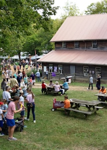 A lively outdoor gathering with numerous people of various ages engaging in activities. The scene includes a rustic wooden building with a metal roof, surrounded by picnic tables where attendees are seated. A group is in line, while others are scattered around the vicinity, possibly part of a fair or festival. Trees and greenery are present, enhancing the natural setting.
