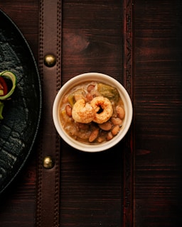 A small white bowl filled with a hearty bean stew containing various ingredients like beans and vegetables, topped with crispy pork rinds. It's placed on a dark wooden surface with a leathery texture and brass accents. To the left, there's a glimpse of a black plate with a garnish of thinly sliced greens and a piece of tomato.