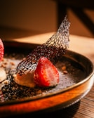 A close-up of a gourmet dessert featuring a fresh halved strawberry on top of a creamy base, garnished with a delicate black mesh-like tuile. The dessert is artistically plated, dusted with powdered sugar, and sprinkled with small crushed nuts on a dark plate.