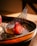 A close-up of a gourmet dessert featuring a fresh halved strawberry on top of a creamy base, garnished with a delicate black mesh-like tuile. The dessert is artistically plated, dusted with powdered sugar, and sprinkled with small crushed nuts on a dark plate.