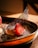 A close-up of a gourmet dessert featuring a fresh halved strawberry on top of a creamy base, garnished with a delicate black mesh-like tuile. The dessert is artistically plated, dusted with powdered sugar, and sprinkled with small crushed nuts on a dark plate.