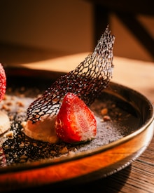A close-up of a gourmet dessert featuring a fresh halved strawberry on top of a creamy base, garnished with a delicate black mesh-like tuile. The dessert is artistically plated, dusted with powdered sugar, and sprinkled with small crushed nuts on a dark plate.