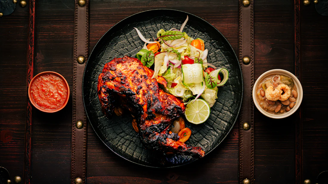 Tandoori chicken on a black plate with salad, lime and chutneys