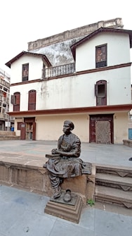 A bronze statue of a seated man wearing traditional clothing, set in front of an old, two-story building with a weathered facade. The structure has closed shutters on its windows and features a rustic architectural style. Stone steps lead up to the statue, and the ground is paved with large tiles.