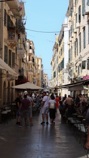 A bustling street scene with narrow passageways flanked by tall, old-style buildings featuring balconies and shutters. People are walking along the street and sitting at outdoor cafes, creating a lively atmosphere. The sky is clear and blue, enhancing the warm, sunny ambiance. Colorful flowers in window boxes add a touch of vibrancy to the setting.