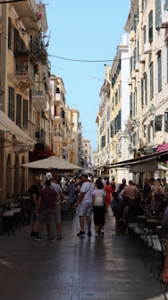 A bustling street scene with narrow passageways flanked by tall, old-style buildings featuring balconies and shutters. People are walking along the street and sitting at outdoor cafes, creating a lively atmosphere. The sky is clear and blue, enhancing the warm, sunny ambiance. Colorful flowers in window boxes add a touch of vibrancy to the setting.