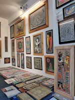 A brightly lit gallery wall displaying a variety of framed traditional paintings, featuring intricate designs and patterns. The artworks are diverse, including depictions of mythological figures, circular motifs, and abstract designs. There is also a stack of similar unframed artworks on the floor below the display.