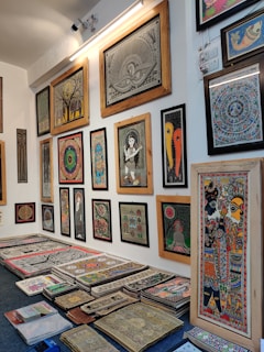 A brightly lit gallery wall displaying a variety of framed traditional paintings, featuring intricate designs and patterns. The artworks are diverse, including depictions of mythological figures, circular motifs, and abstract designs. There is also a stack of similar unframed artworks on the floor below the display.