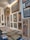 A brightly lit gallery wall displaying a variety of framed traditional paintings, featuring intricate designs and patterns. The artworks are diverse, including depictions of mythological figures, circular motifs, and abstract designs. There is also a stack of similar unframed artworks on the floor below the display.