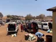 A busy parking lot filled with several vans and people loading or unloading goods. Some people are bending over bags and sacks, while others are standing near the vehicles. The area appears to be urban, with buildings and trees in the background under a clear blue sky.