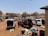 A busy parking lot filled with several vans and people loading or unloading goods. Some people are bending over bags and sacks, while others are standing near the vehicles. The area appears to be urban, with buildings and trees in the background under a clear blue sky.