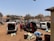 A busy parking lot filled with several vans and people loading or unloading goods. Some people are bending over bags and sacks, while others are standing near the vehicles. The area appears to be urban, with buildings and trees in the background under a clear blue sky.