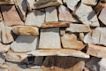 An arrangement of natural stone slabs stacked in a seemingly random pattern, creating a rustic and textured surface. The stones vary in size and shape, with a mix of flat and irregular edges. They exhibit a range of earth tones, including light beige, brown, and hints of reddish hues. The surface displays natural imperfections and variations in color, highlighting the organic appearance of the stone material.