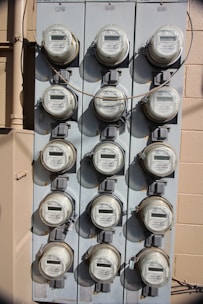 A vertical, metal panel mounted on a wall houses twelve circular electricity meters. Each meter is installed in a row and column arrangement, with cables running across and connecting them. The wall behind the panel is beige with visible shadows.