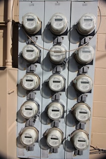 A vertical, metal panel mounted on a wall houses twelve circular electricity meters. Each meter is installed in a row and column arrangement, with cables running across and connecting them. The wall behind the panel is beige with visible shadows.
