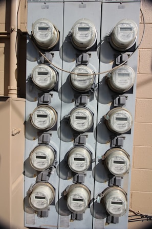 A vertical, metal panel mounted on a wall houses twelve circular electricity meters. Each meter is installed in a row and column arrangement, with cables running across and connecting them. The wall behind the panel is beige with visible shadows.