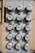A vertical, metal panel mounted on a wall houses twelve circular electricity meters. Each meter is installed in a row and column arrangement, with cables running across and connecting them. The wall behind the panel is beige with visible shadows.