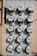 A vertical, metal panel mounted on a wall houses twelve circular electricity meters. Each meter is installed in a row and column arrangement, with cables running across and connecting them. The wall behind the panel is beige with visible shadows.