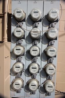 A vertical, metal panel mounted on a wall houses twelve circular electricity meters. Each meter is installed in a row and column arrangement, with cables running across and connecting them. The wall behind the panel is beige with visible shadows.