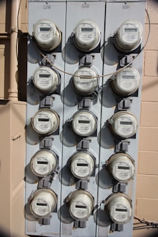 A vertical, metal panel mounted on a wall houses twelve circular electricity meters. Each meter is installed in a row and column arrangement, with cables running across and connecting them. The wall behind the panel is beige with visible shadows.