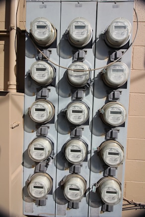 A vertical, metal panel mounted on a wall houses twelve circular electricity meters. Each meter is installed in a row and column arrangement, with cables running across and connecting them. The wall behind the panel is beige with visible shadows.