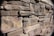 A close-up view of a stone wall made up of variously sized, horizontally stacked stones. The stones have a rough texture and display a range of earth tones, including grays, browns, and hints of reddish hues. Shadows between the stones indicate a natural, uneven surface.