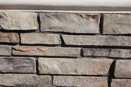 A close-up view of a stone wall constructed with rectangular stones arranged in a horizontal pattern. The stones vary in shades of grey and brown with subtle texture differences, displaying natural imperfections.