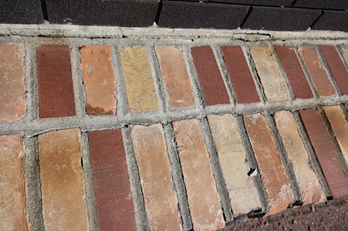 A close-up view of a brick wall section with rectangular bricks arranged in a pattern, featuring varied shades of red and beige. The mortar between the bricks is visible, showing signs of wear and uneven surfaces.