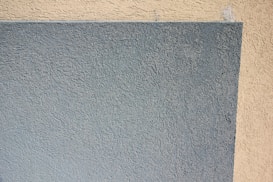 A textured wall surface with a rectangular section in two colors. The left side of the wall is painted a muted blue-gray, while the adjacent section is a light beige. The texture is rough with a stucco-like finish.