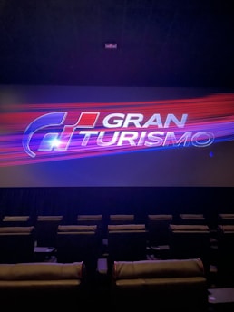 A theater setting with empty reclining seats arranged in rows, facing a large screen. On the screen, there's an image with the text 'GRAN TURISMO' overlaying streaks of blue and red light.