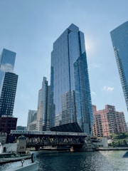 A cityscape featuring tall modern skyscrapers with reflective glass facades, a bridge spanning a body of water, and a train crossing the bridge. The sunlight reflects off the buildings, contributing to a bright, clear sky.