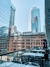 A cityscape featuring modern high-rise buildings with glass facades and a historic building labeled 'Hudson's Bay.' In the foreground, a rooftop patio is visible with tables and chairs, some of which are occupied by people enjoying a meal.