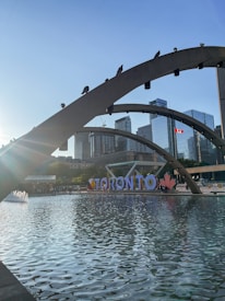 An urban scene featuring a large reflective pool with modern arched bridges. In the background, high-rise buildings create a cityscape, complemented by a Canadian flag fluttering in the wind. The word 'TORONTO' is prominently displayed along the waterfront accompanied by a red maple leaf symbol. The sun casts a bright flare from the left, adding a dynamic element to the composition.